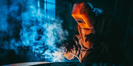 Welder in protective gear works on welding steel, surrounded by hazardous fumes from the process.