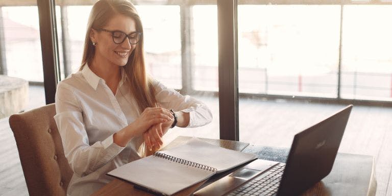A manager checking her watch whilst sat working at a desk with a laptop and notebook