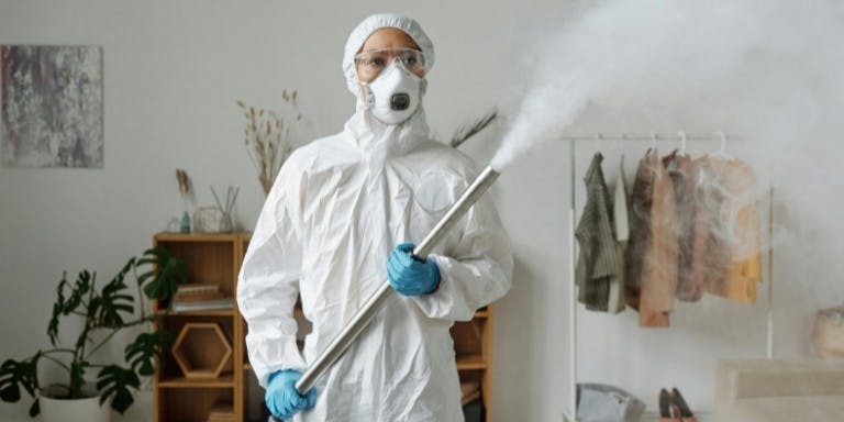 Person in in a protective suit and mask sprays disinfectant in a room, demonstrating COSHH PPE safety measures.