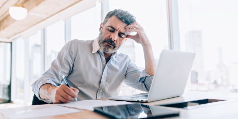 Male small business owner calculating weekly rota cost from wage increase at a desk with laptop, paper and tablet.