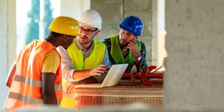 Staggered work hours Three construction workers in hard hats and vests collaborate over a tablet, discussing staggered work hours on site.
