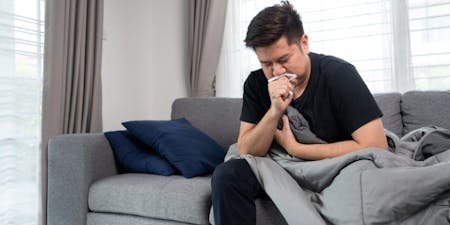 Managing sickness at work Man on couch with a blanket coughing into a tissue.