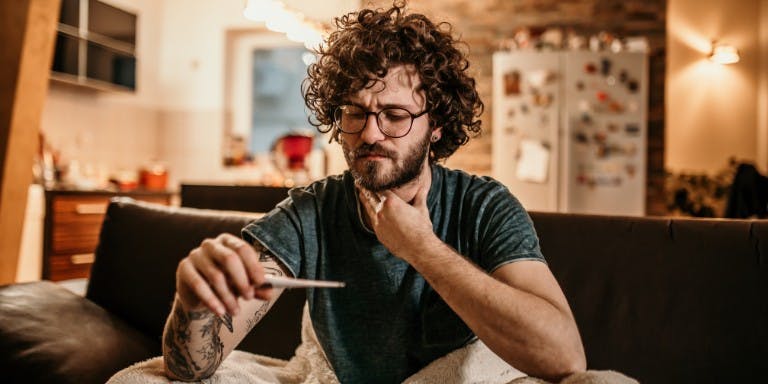 Contractual sick pay Man with curly hair on a couch checking temperature, showing signs of illness and fatigue.
