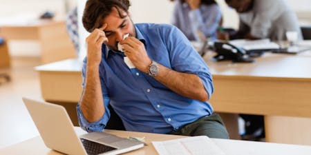 Contractual sick pay Man at a desk with his head in his hands, appearing unwell and stressed.
