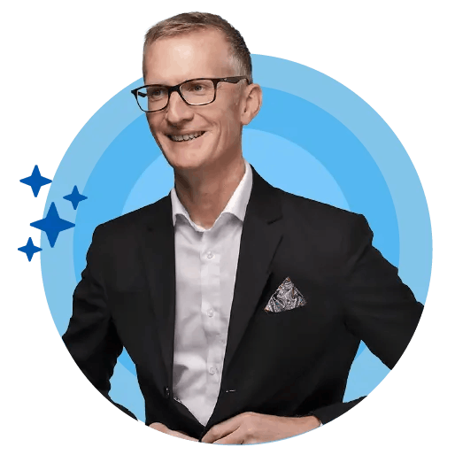 A man with glasses and a black suit, wearing a white shirt and a pink pocket square, is smiling and looking directly at the camera. The background features a blue circle with stars, and the man is positioned centrally within the circle. A man with glasses and a black suit, wearing a white shirt and a pink pocket square, is smiling and looking directly at the camera. The background features a blue circle with stars, and the man is positioned centrally within the circle.