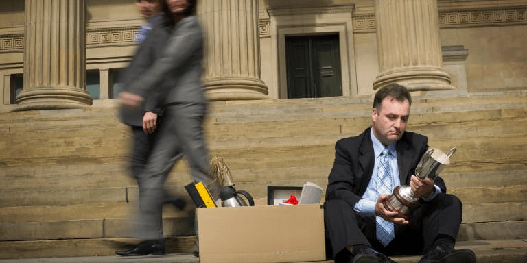 A man made redundant in a black suit sits on a set of steps, holding a silver cup and a silver cup holder. Behind him, a woman walks past a building with black doors and a black window.