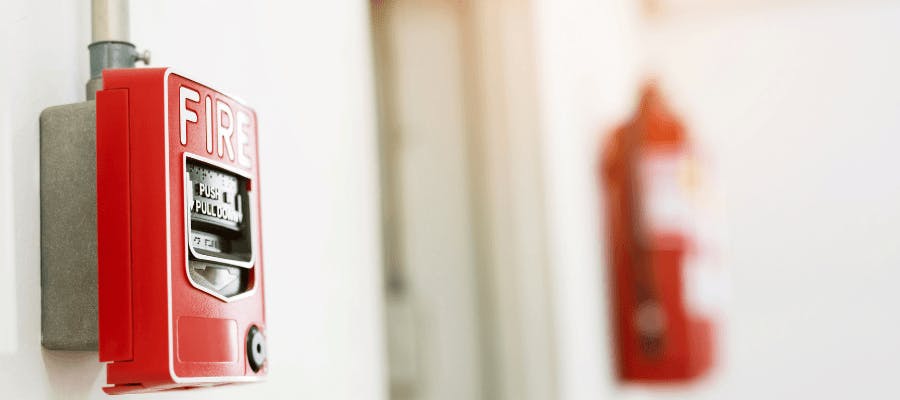 A red fire hydrant is attached to a white wall, with a gray fire hose running vertically on the right side. The fire hydrant has a white label with black text that reads "FIRE HAZARD" and "1000". The fire hose is positioned to the right of the fire hydrant.