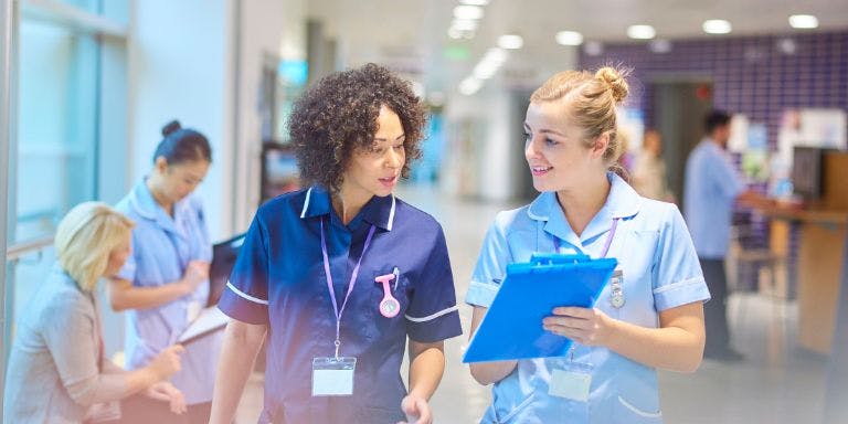 Two nurses are standing in a hallway, engaged in a conversation. One nurse is holding a clipboard, while the other nurse is holding a binder. They both appear to be in a professional setting, possibly a hospital or a nursing facility. Two nurses are standing in a hallway, engaged in a conversation. One nurse is holding a clipboard, while the other nurse is holding a binder. They both appear to be in a professional setting, possibly a hospital or a nursing facility.