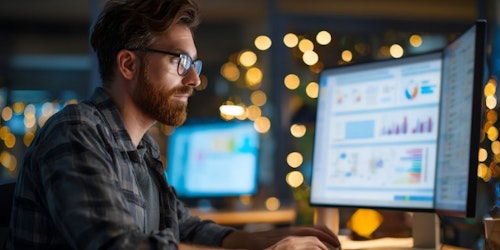 A man with glasses and a beard is sitting at a desk, working on a computer. The desk is surrounded by three computer monitors, each displaying a different graph. The man is focused on the screen, indicating that he is engaged in some form of data analysis or research. The background features a window with lights, adding a warm and cozy atmosphere to the scene.