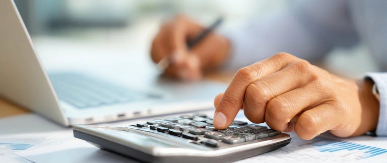 A person's hand is typing on a silver laptop computer, with a pen in their hand. The laptop is on a desk with blue financial documents. The background is blurred, but it appears to be a workspace or office setting.