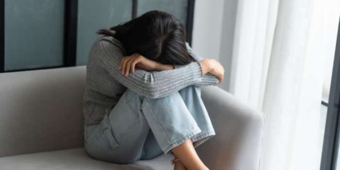 A woman is sitting on a gray couch, her head resting on her hand. She is wearing a gray sweater and blue jeans. The background features a window with white curtains.