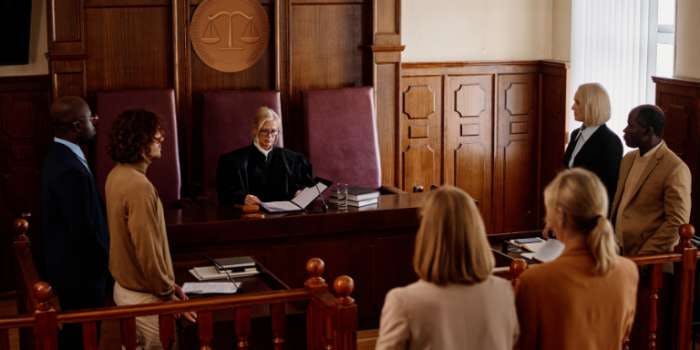 In the image, a woman is seated at a wooden desk in a courtroom. She is wearing a suit and is surrounded by several people. The woman is facing a judge who is seated at the desk. The judge is wearing a suit and is holding a document. The people around the desk are engaged in a discussion, with one person holding a pen. The background features a wooden wall and a window.