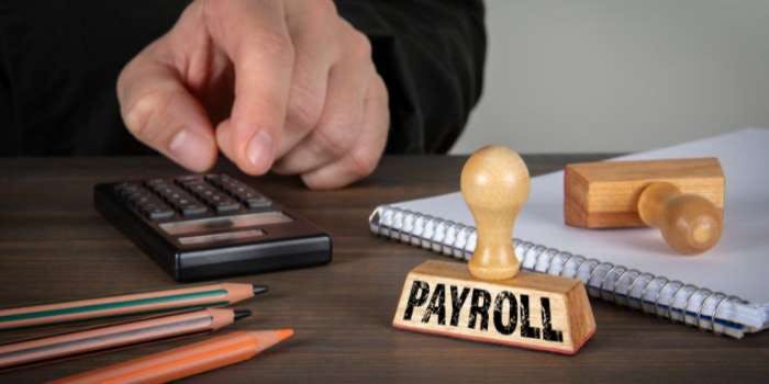 A person is sitting at a desk with various office supplies and a calculator on it. The person is holding a key and is looking at a "paycheck" sign. The desk also has a binder, a notebook, and a pen.