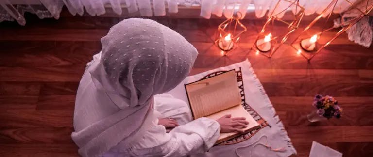A person in a white headscarf and prayer hat is sitting on a wooden floor, engrossed in reading a book. The book is open and resting on a white rug. The room is illuminated by a series of lit candles, casting a warm glow. A vase with flowers is placed to the side of the person. The person's position and the book's orientation suggest a quiet, contemplative moment.