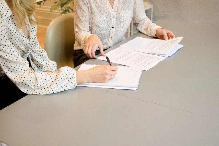 Two individuals are seated at a gray desk, engaged in a conversation. The person on the left is holding a pen and writing on a document, while the person on the right is holding a pen and also writing on a document. The desk is cluttered with papers and documents, suggesting a work environment. In the background, there's a plant and a window, providing a natural and calming backdrop to the scene.
