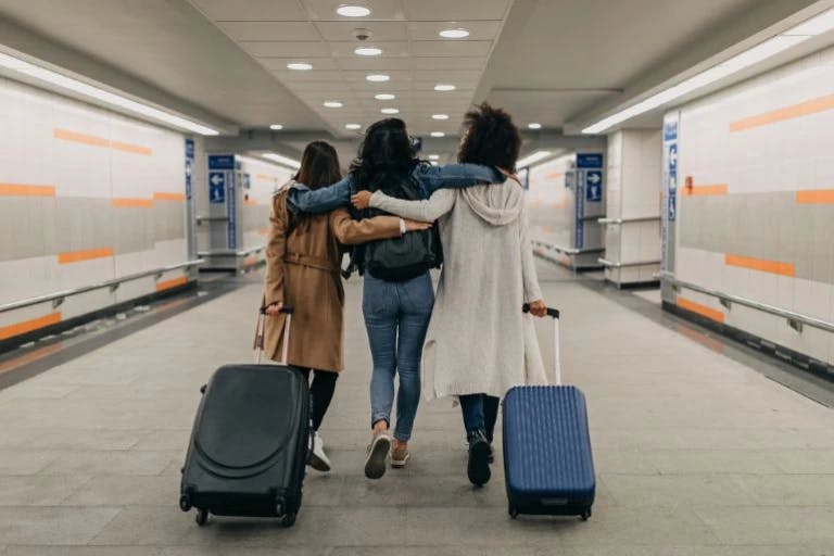 Three women are walking together in an airport terminal. They are hugging each other and are carrying luggage. The terminal has white and orange tiles on the walls. The woman on the left is wearing a white coat, the middle woman is wearing a brown coat, and the woman on the right is wearing a blue coat. They are moving towards the right side of the image.