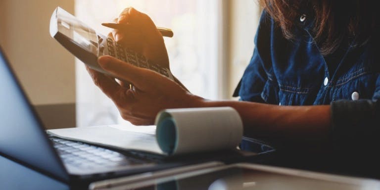 A person is sitting at a desk, holding a pen and a piece of paper. The desk has a laptop and a notebook on it. The person is wearing a blue shirt. The background shows a window with a view of a street.