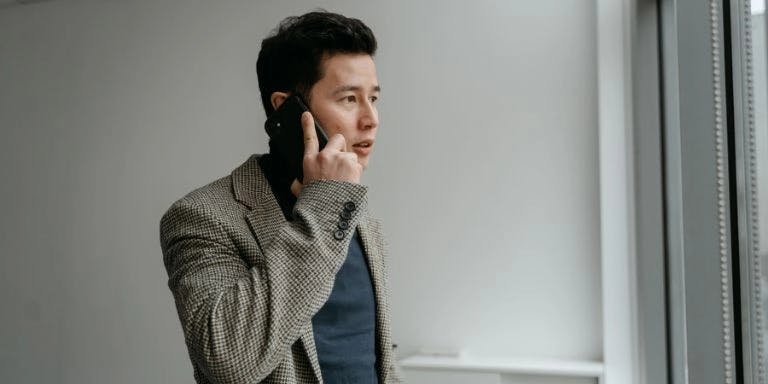 A man is standing in front of a window, holding a phone to his ear, and looking off to the side. He is dressed in a gray blazer and a blue shirt. The window behind him has blinds.