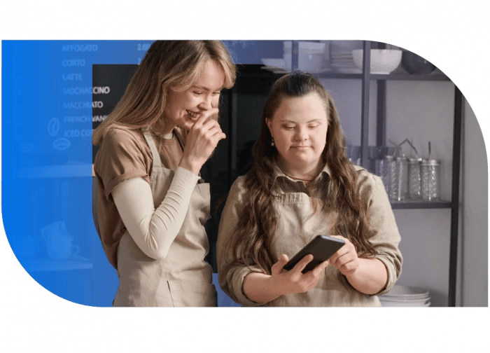 In the image, there are two women standing in front of a blue wall. The woman on the left is wearing a beige shirt and has long brown hair. She is looking at a tablet, possibly reading or interacting with something on it. The woman on the right is wearing a light-colored shirt and has long brown hair as well. She is also looking at the tablet and seems to be in a conversation or discussion. Both women are focused on the tablet, indicating a moment of interaction or engagement.