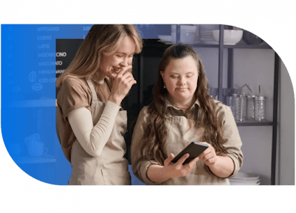 In the image, there are two women standing in front of a blue wall. The woman on the left is wearing a beige shirt and has long brown hair. She is looking at a tablet, possibly reading or interacting with something on it. The woman on the right is wearing a light-colored shirt and has long brown hair as well. She is also looking at the tablet and seems to be in a conversation or discussion. Both women are focused on the tablet, indicating a moment of interaction or engagement.