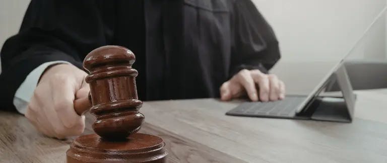 A judge's robe, a wooden gavel, and a laptop computer are in the image. The gavel is on a wooden table, and the laptop is on a desk. The judge is seated at the table, and the laptop is on the desk. The background is blurred, but it appears to be a white wall.