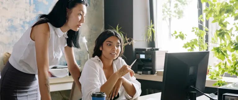 In the image, there are two women in an office setting. One woman is seated at a desk, holding a pen and a pencil, and appears to be working on a computer. The other woman is standing behind the desk, holding a phone and looking at the computer screen. The desk is cluttered with various objects, including a computer monitor, a printer, and a soda can. The office has a window with a plant in the background, and the wall has a blue wallpaper. The woman