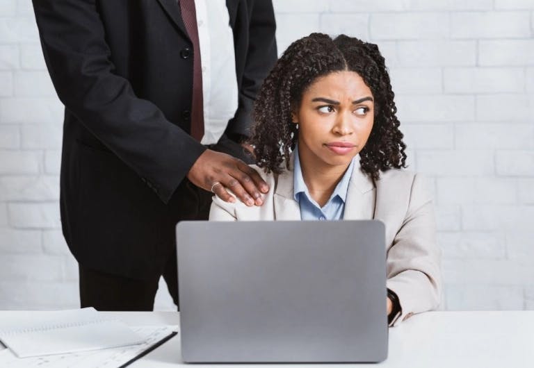 A woman in a beige blazer and blue shirt is seated at a white desk, looking serious and focused on her laptop. Behind her, a man in a black suit is leaning over her, his hand resting on her shoulder. The desk is cluttered with papers and a laptop, suggesting a work environment. The background is a white brick wall, providing a neutral backdrop that emphasizes the woman and the man. The image captures a moment of intense concentration and professionalism.