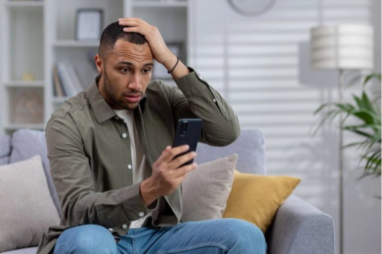 A man in a green shirt and blue jeans is sitting on a gray couch in a living room. He is holding a black phone in his right hand and his left hand is on his head. The room has a white wall, a bookshelf, and a lamp. The man appears to be in a moment of concern or frustration.