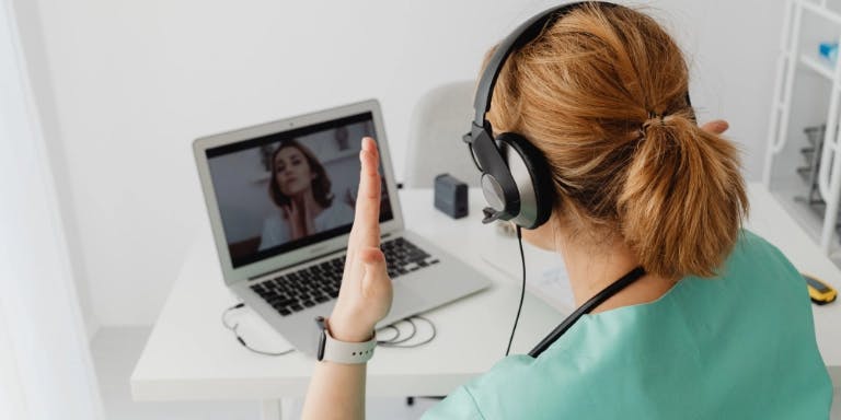 A person wearing a green scrubs is sitting at a desk, using a laptop and a microphone. The person is wearing a pair of headphones and has a microphone in their hand. The desk has a white surface and a black computer mouse. The person is pointing towards the microphone, indicating they are engaged in a conversation. A person wearing a green scrubs is sitting at a desk, using a laptop and a microphone. The person is wearing a pair of headphones and has a microphone in their hand. The desk has a white surface and a black computer mouse. The person is pointing towards the microphone, indicating they are engaged in a conversation.