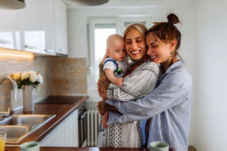 In a cozy kitchen, a mother and her baby are sharing a warm moment. The mother, dressed in a striped shirt, is holding her baby in her arms. The baby, dressed in a blue and white polka dot shirt, is looking at the camera with a smile. The kitchen is well-lit, with white cabinets and a sink visible in the background. A vase of flowers adds a touch of nature to the scene. The mother and baby are standing near the counter, which has a In a cozy kitchen, a mother and her baby are sharing a warm moment. The mother, dressed in a striped shirt, is holding her baby in her arms. The baby, dressed in a blue and white polka dot shirt, is looking at the camera with a smile. The kitchen is well-lit, with white cabinets and a sink visible in the background. A vase of flowers adds a touch of nature to the scene. The mother and baby are standing near the counter, which has a