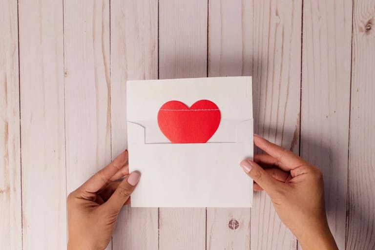 A pair of hands is holding a white envelope with a red heart on it, placed on a wooden surface. The envelope is open, revealing the red heart. The background is a light-colored wooden surface with a small hole.