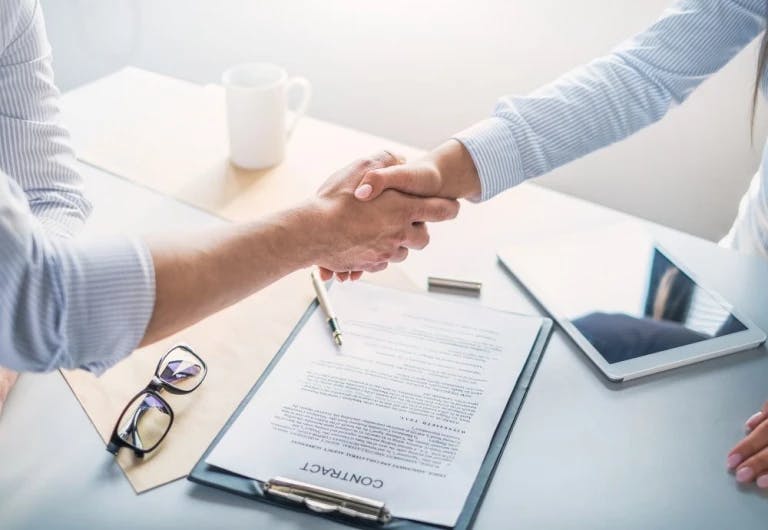 Two individuals are in a professional setting, with one person shaking hands over a document. The desk is cluttered with various objects, including a laptop, a pen, a notebook, and a mug. The person on the left is wearing a white shirt, while the person on the right is in a blue shirt. The background is blurred, focusing on the main subjects.