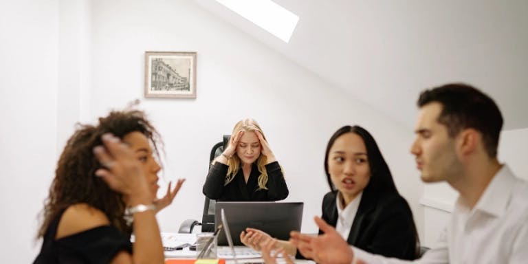In the image, there are three individuals seated around a table in an office setting. The person on the left is holding a phone to their ear, indicating they are engaged in a conversation. The middle person is gesturing with their hand, possibly explaining something to the other two. The person on the right is gesturing with their hand, possibly responding to the middle person's question or providing feedback. The table is cluttered with papers and a laptop, suggesting a productive meeting. The background features a