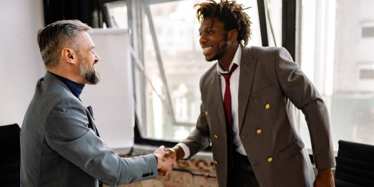 Two men are shaking hands in an office setting. One man is dressed in a gray suit with a red tie, while the other man is wearing a blue shirt and a black tie. They are positioned in front of a whiteboard and a window with a view of a cityscape. The man on the left has a beard and is wearing a blue shirt, while the man on the right has curly hair and is wearing a gray suit with a red tie.