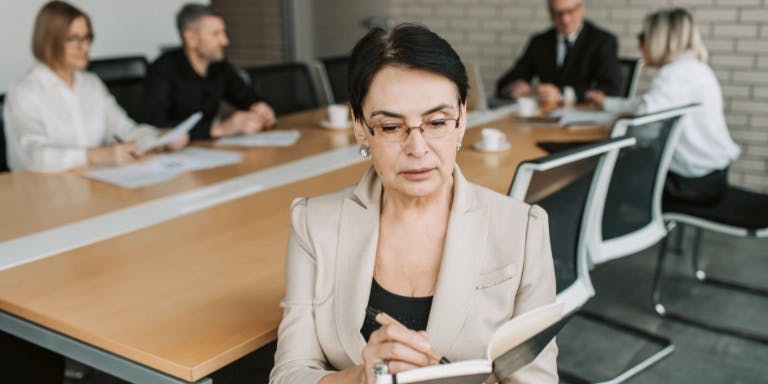 A woman in a beige blazer is seated at a wooden conference table, engrossed in a document. Behind her, three men are seated at a separate table, each holding a laptop. The room has a brick wall and a window, providing a contrast to the modern office setting. The woman's focused expression and the men's focused laptops suggest a serious meeting or discussion.