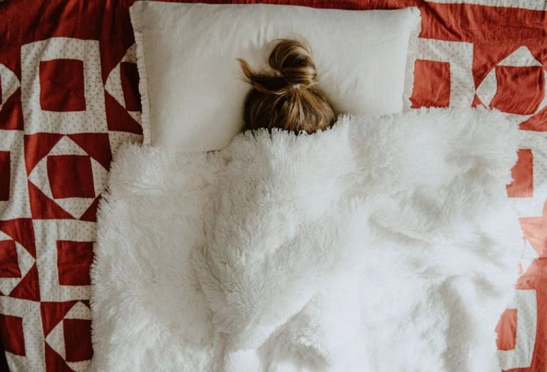 A person is lying on a bed covered with a white blanket and a red and white quilt. The person's hair is tied up in a bun.