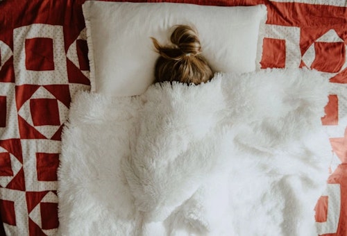 A person is lying on a bed covered with a white blanket and a red and white quilt. The person's hair is tied up in a bun.