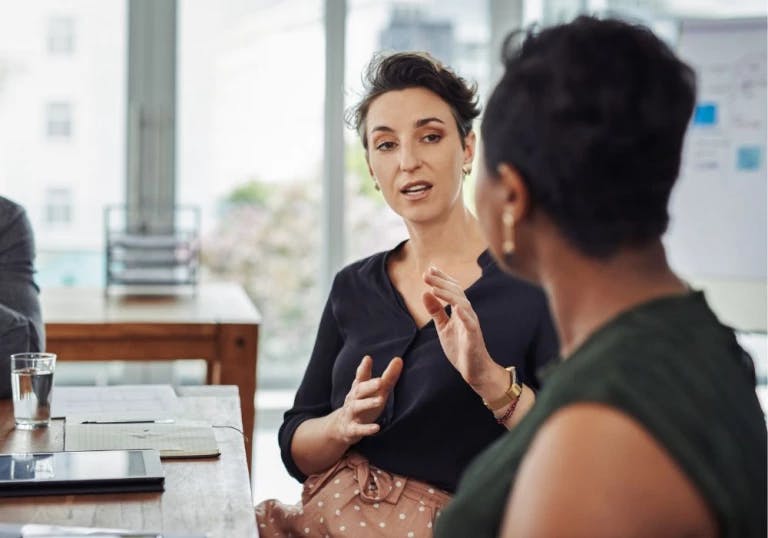 In the image, a woman is seated at a desk in a modern office setting. She is dressed in a black top and a pink skirt, and her hands are raised in a gesture of discussion. Behind her, another woman is seated at a different desk, also dressed in a black top and a pink skirt. They are both engaged in a conversation, with the woman in the black top speaking to the woman in the pink skirt. The desk they are sitting at has a laptop and a glass of