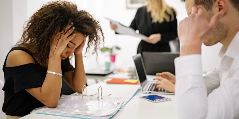 A woman with curly hair is sitting at a desk, holding her head over her eyes, and appears to be in a state of distress. She is wearing a black top and has a white bracelet on her wrist. In front of her, there is a blue notebook and a white pen. To her right, there is a man who is holding a blue laptop. In the background, there is a woman who is holding a white clipboard. The desk is cluttered with papers and a notebook.