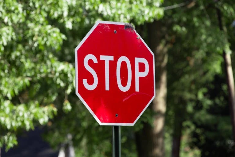 A red stop sign with the word "STOP" written in white is prominently displayed in the foreground. The sign is attached to a green pole, which is slightly tilted to the left. The background features lush green trees and bushes, providing a natural backdrop to the sign. The image does not contain any other objects or text. A red stop sign with the word "STOP" written in white is prominently displayed in the foreground. The sign is attached to a green pole, which is slightly tilted to the left. The background features lush green trees and bushes, providing a natural backdrop to the sign. The image does not contain any other objects or text.