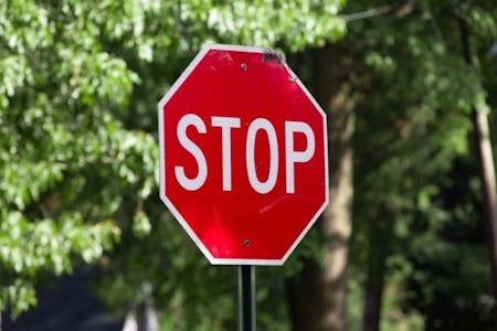 A red stop sign with the word "STOP" written in white is prominently displayed in the foreground. The sign is attached to a green pole, which is slightly tilted to the left. The background features lush green trees and bushes, providing a natural backdrop to the sign. The image does not contain any other objects or text.