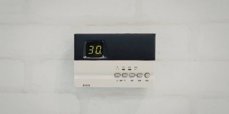 A white wall with a black border displays a digital thermostat. The digital display shows the time as 12:05. The thermostat is mounted on the wall, with a label "1205" visible.