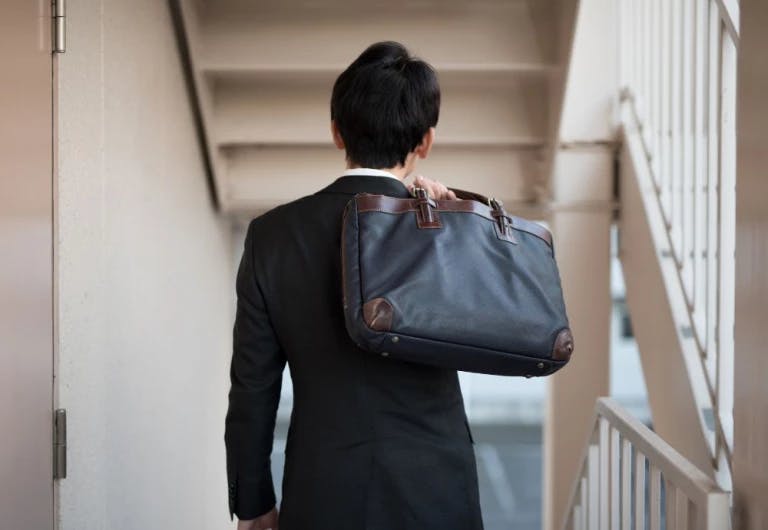 A man in a black suit is walking down a hallway, carrying a large blue leather messenger bag with brown straps. The man's hair is neatly tied up in a bun. The hallway has white walls and a white staircase with a brown railing. The man appears to be heading towards a building.