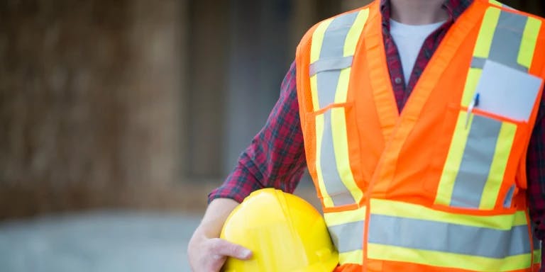 A person wearing a yellow safety vest and a yellow hard hat is standing in front of a building with a wooden wall. The person's hands are clasped together, and the background is blurred, focusing on the person and the building.