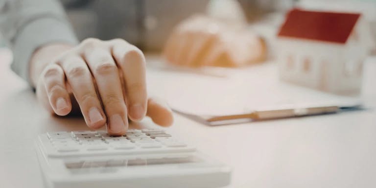A hand is typing on a white calculator, with a pen resting on the table. A small white house with a red roof is visible in the background. The scene suggests a workspace or home office setting.