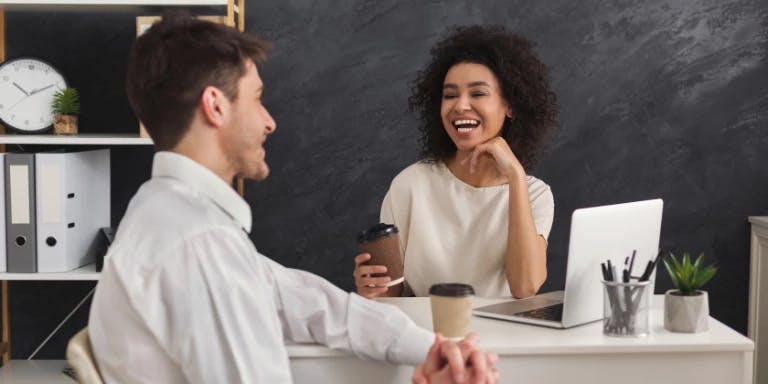 A man and a woman are in an office setting. The man is sitting at a desk, holding a coffee cup and smiling. The woman is seated next to him, holding a phone and smiling as well. The desk has a laptop, a mug, a clock, and a plant. The background features a blackboard with a clock and a plant.