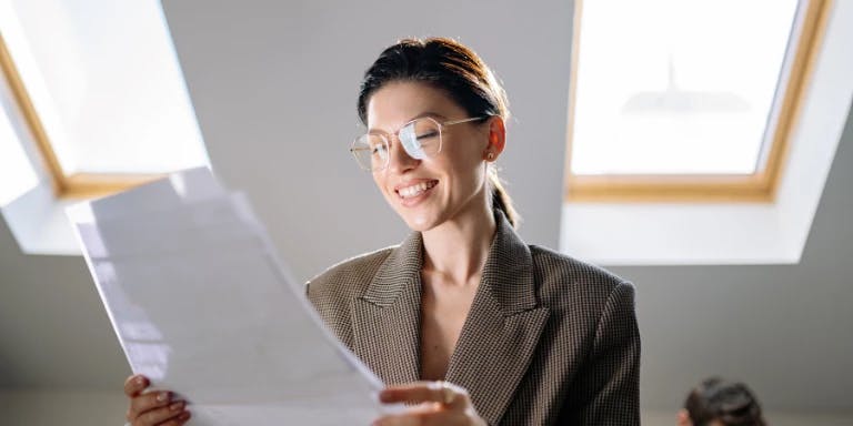 A woman is seated in a room with two windows, one on each side of her. She is wearing glasses and a brown blazer. She is holding a piece of paper in her hands and appears to be looking at it. The room has a white wall and a wooden desk in front of her.