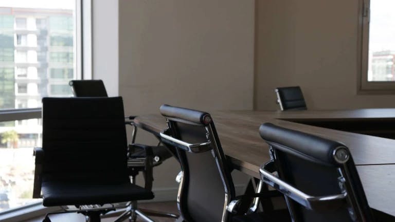 The image depicts a well-lit conference room with a wooden table in the foreground. Six black office chairs are arranged around the table, facing the table. The room has a minimalist aesthetic with white walls and a large window on the right side. The floor is made of wood, and the room appears to be empty, with no people visible in the image.