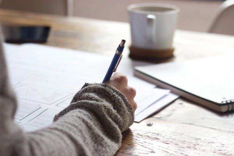 A person is sitting at a wooden table, holding a blue pen and writing on a piece of paper. A white mug is on the table, and a notebook is nearby. The person is wearing a sweater. The background is blurred, but it appears to be a room with a window.