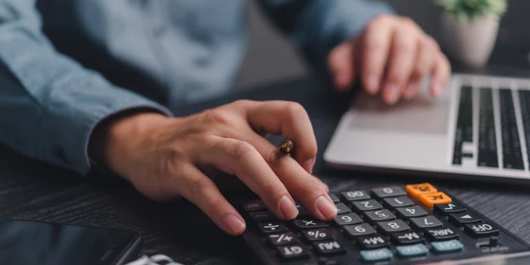 A person is typing on a black calculator with a blue and orange button, while a laptop is open on a desk. A potted plant is in the background.